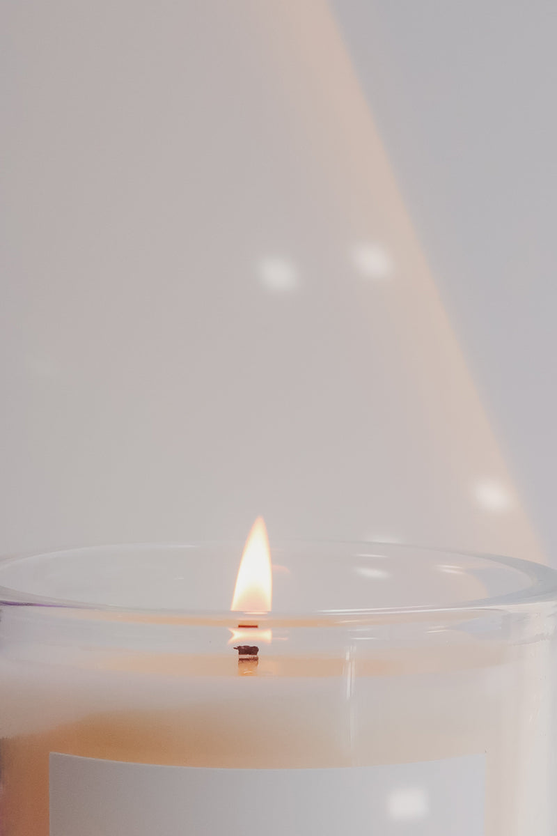 A lit candle in a clear glass jar on a light grey background with a sunray reflection on the background. 