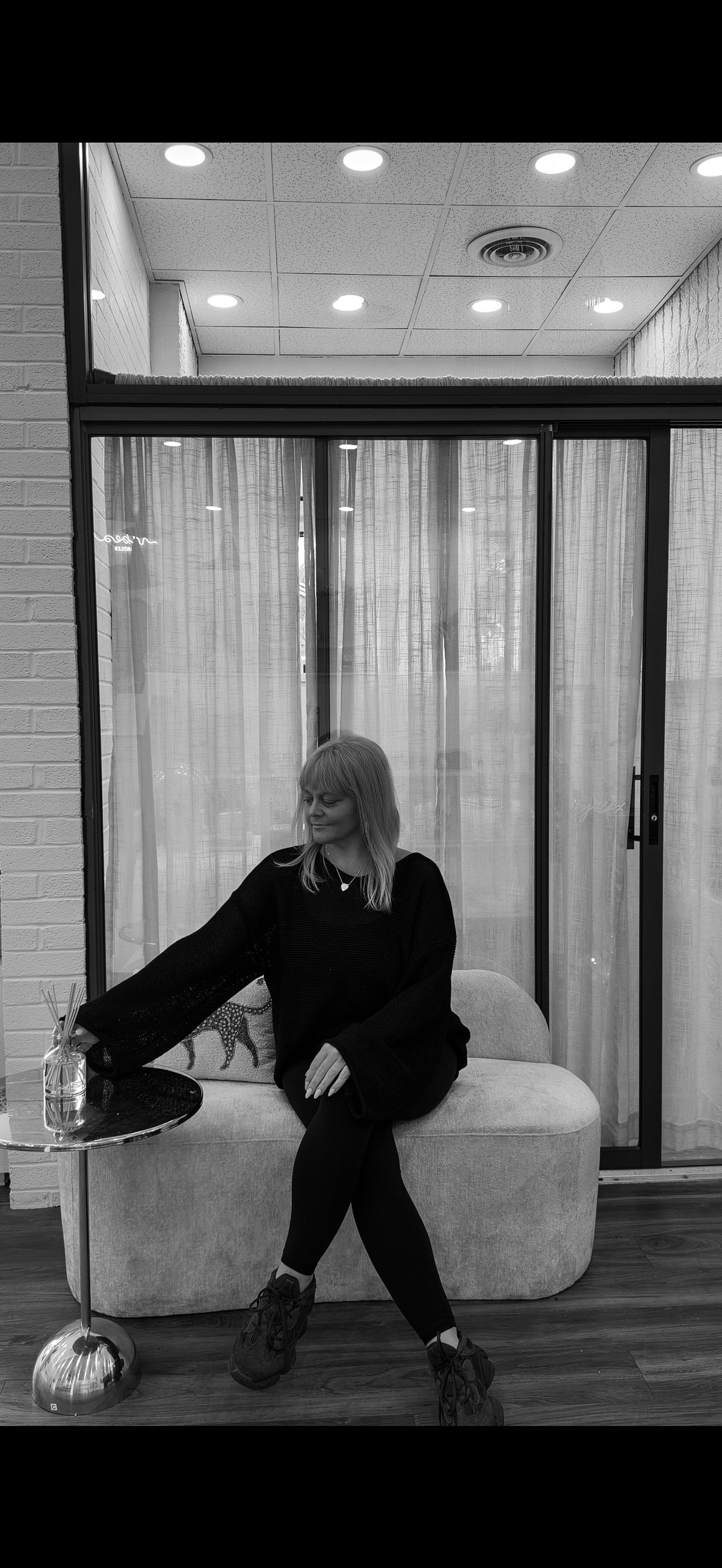 A woman sits on a light-colored sofa in a modern room with large glass doors and curtains, wearing a black long-sleeve top, black pants, and dark sneakers. The photo is in black and white.